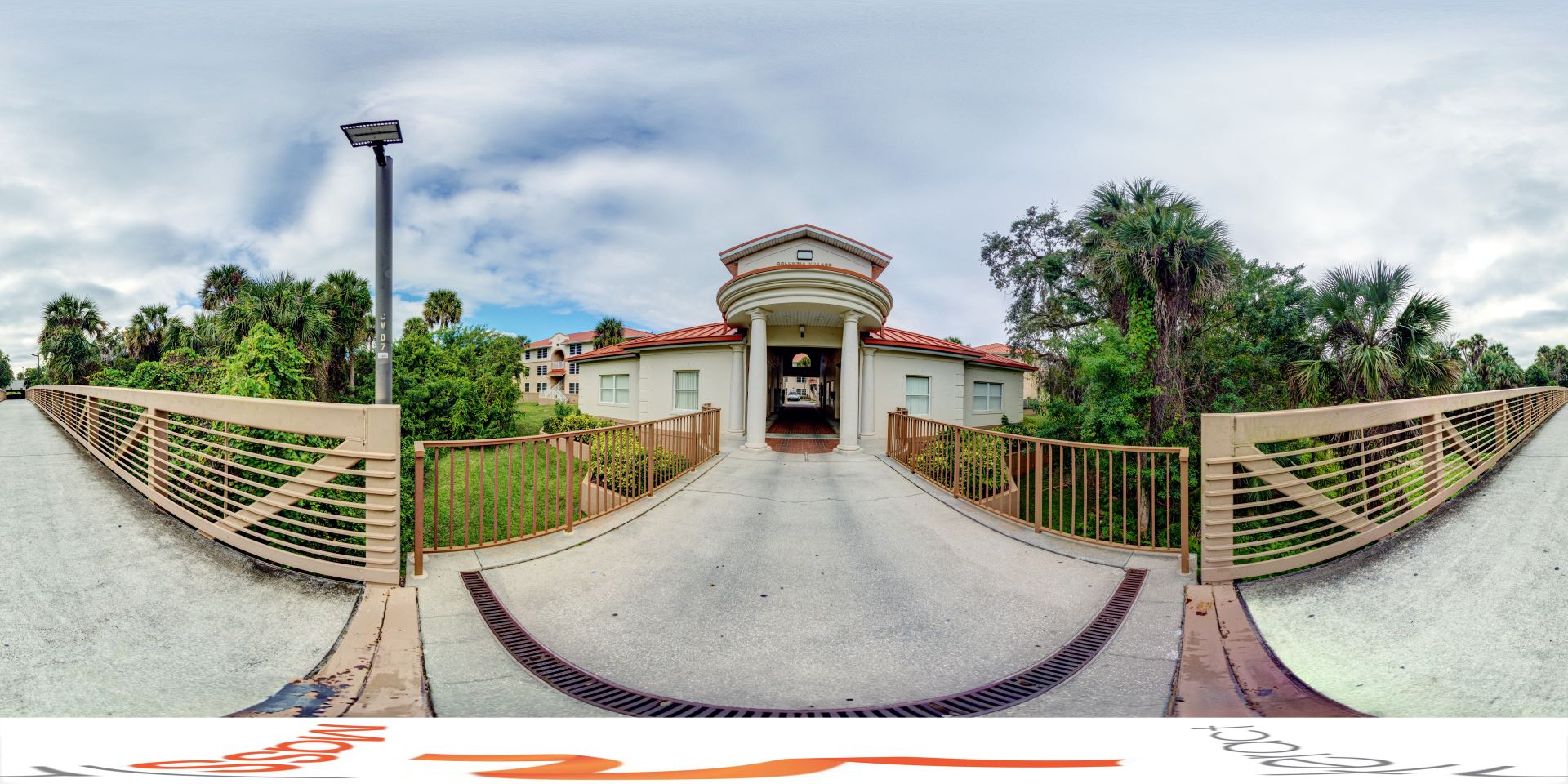 Panoramic view of a walkway leading to a building with columns, surrounded by greenery and palm trees, under a cloudy sky